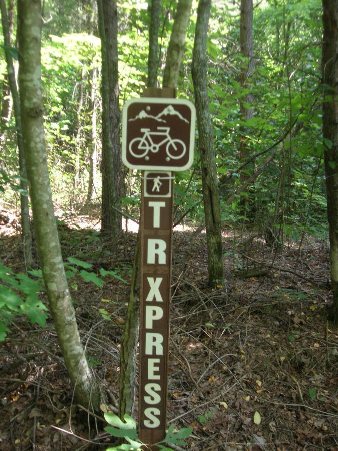 A trail sign indicating a bike path, featuring a bicycle symbol and the word "TRX PRESS," surrounded by trees and greenery in a forested area. Tanasi Trail System mountain bike trail.