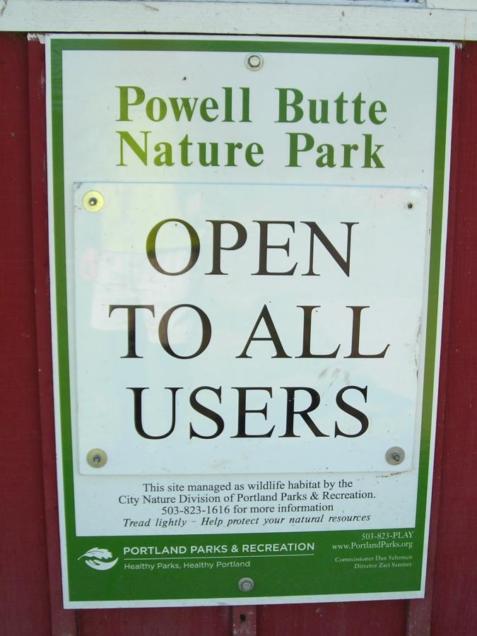 Sign for Powell Butte Nature Park stating "OPEN TO ALL USERS." The sign includes information about the park being managed as a wildlife habitat by the City Nature Division of Portland Parks & Recreation, along with a contact number for more information. The bottom portion features the logos of Portland Parks & Recreation. Powell Butte Nature Park mountain bike trail.