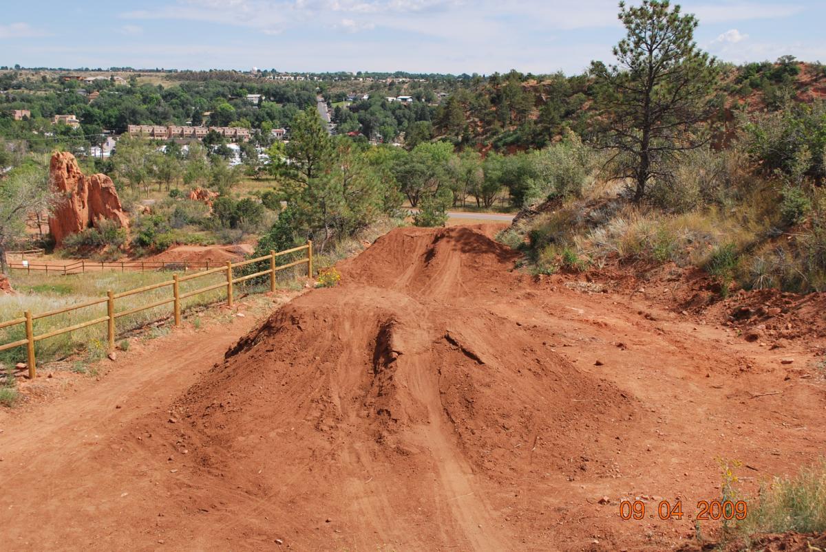 A dirt bike track featuring two large jumps surrounded by a natural landscape of greenery and rocky formations, with a distant view of a town nestled between hills under a partly cloudy sky. A wooden fence runs alongside the track, indicating a designated area for biking activities. Red Rock Canyon mountain bike trail.