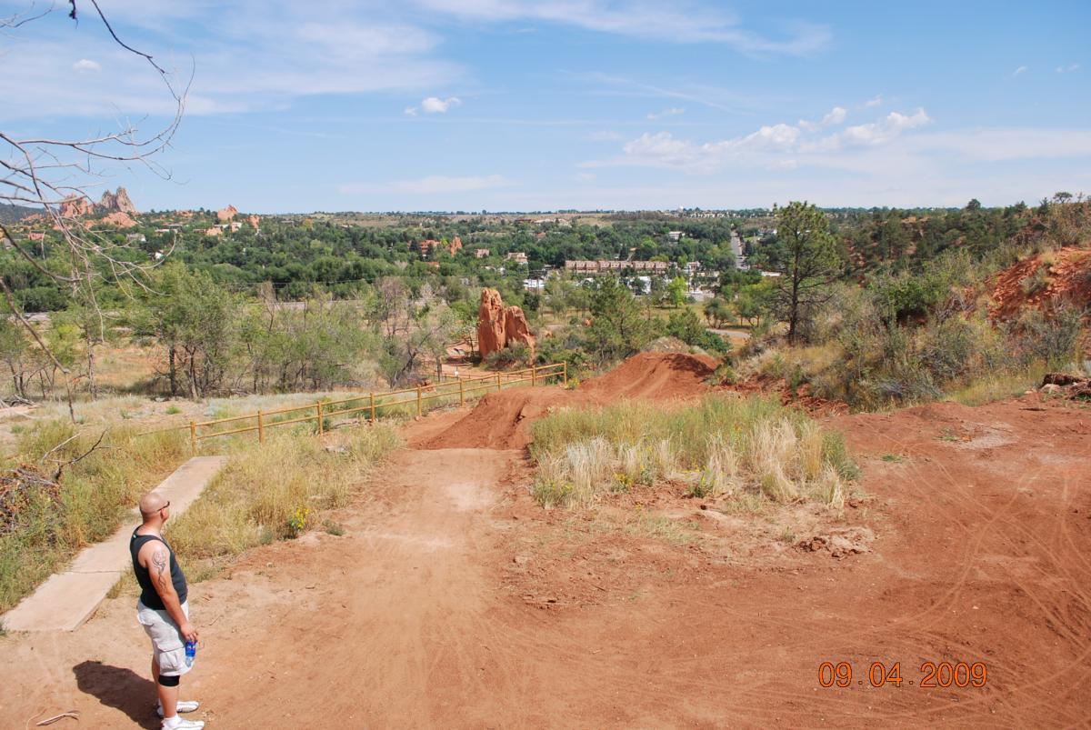 A man stands on a dirt path overlooking a scenic view of green trees and rocky formations in the distance, under a blue sky with scattered clouds. The foreground features dry grass and a dirt track, with a bench along the path. The date in the corner reads September 4, 2009. Red Rock Canyon mountain bike trail.