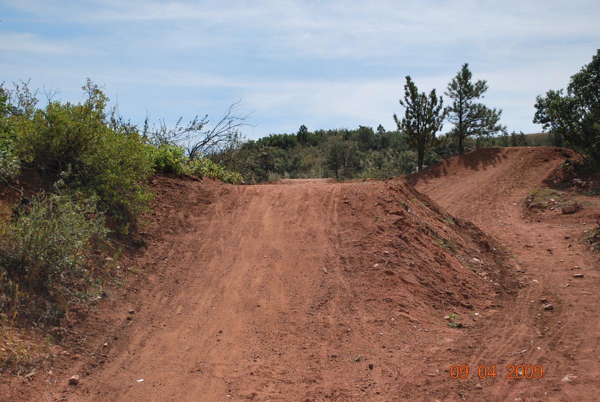 A dirt trail winding through a natural landscape, flanked by shrubs and small trees, under a clear blue sky. The path curves to the right, creating a visible divide between two sections of the terrain. The image captures a sunny outdoors scene, suggesting a recreational area for hiking or biking. The date at the bottom indicates it was taken on September 4, 2009. Red Rock Canyon mountain bike trail.