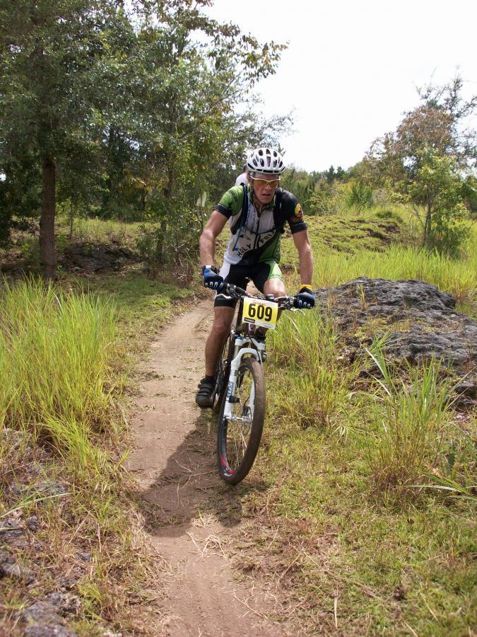 A person riding a mountain bike on a narrow dirt trail surrounded by greenery. The cyclist wears a helmet, gloves, and a numbered racing vest, and is navigating a curved path near some rocks. The sun is shining, indicating a clear day. Haile's Trails mountain bike trail.