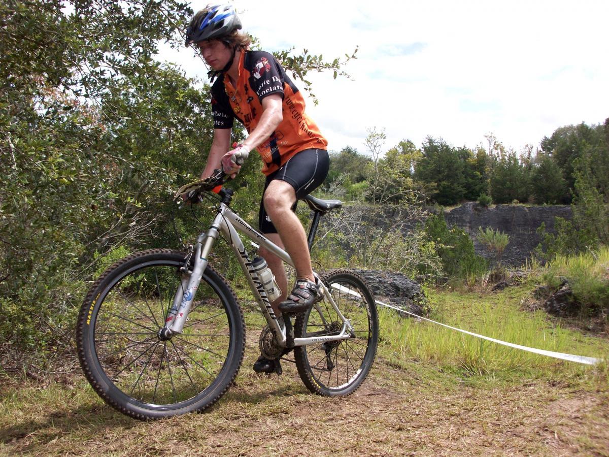 A young cyclist in an orange and black jersey rides a mountain bike along a grassy trail surrounded by trees. The rider is focused, with a slight lean forward, navigating a rocky area in the background. Haile's Trails mountain bike trail.