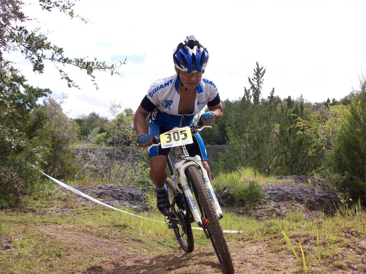 A mountain biker in a blue and white jersey, wearing a helmet, navigates a rugged trail surrounded by trees. The racer, numbered 305, is leaning forward on the bike, demonstrating speed and focus as they tackle the terrain. Haile's Trails mountain bike trail.