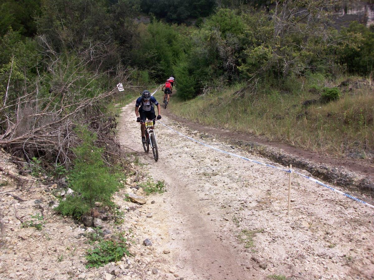 Two mountain bikers navigate a rugged dirt trail surrounded by sparse vegetation and trees. The path is uneven and marked by rocks, with a few sticks and signs indicating race boundaries. The biker in the foreground is leaning forward, focused on maneuvering, while the second biker is visible further up the trail. Haile's Trails mountain bike trail.