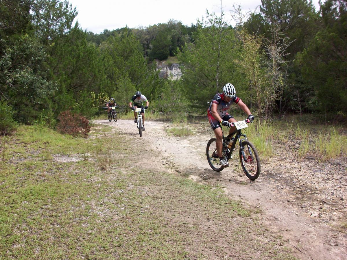 Three mountain bikers racing on a dirt path surrounded by greenery. The riders are focused and are wearing helmets and cycling gear. The terrain includes patches of grass and gravel, and the background features trees and a rocky incline. Haile's Trails mountain bike trail.