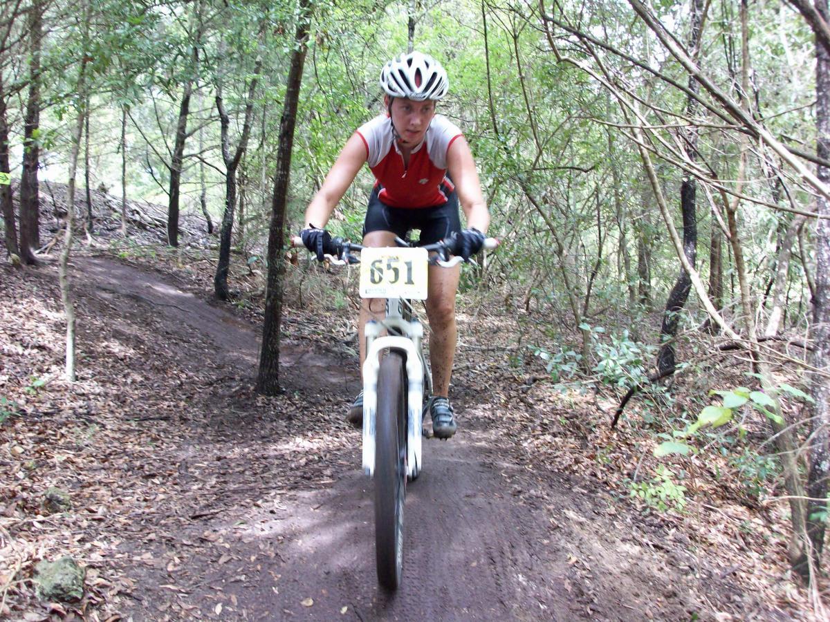 A cyclist in a red and black outfit rides a mountain bike on a dirt trail surrounded by trees. The bike has a race number displayed on it. The environment is wooded, with a mix of sunlight and shadows filtering through the leaves. Haile's Trails mountain bike trail.