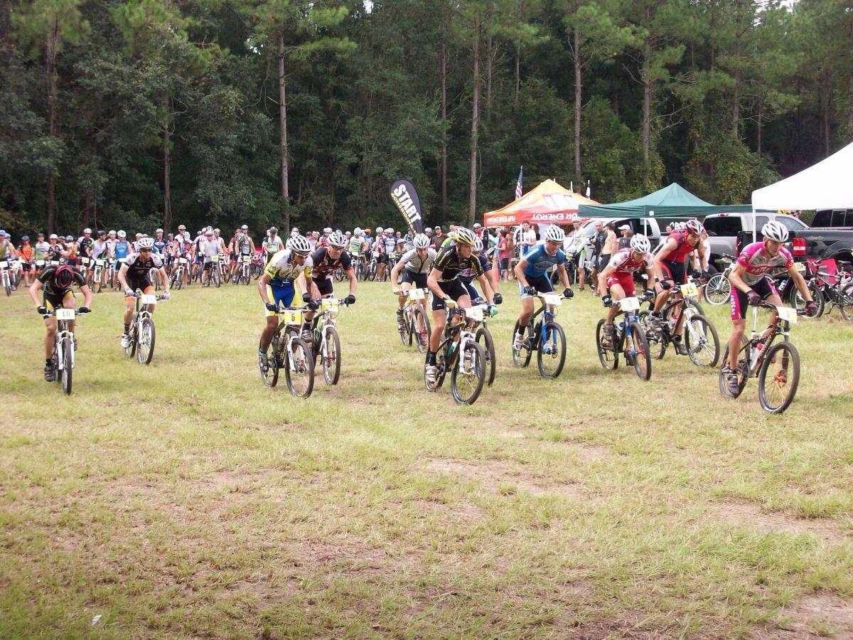 A group of mountain bikers racing on a grassy field, with spectators in the background. The cyclists are wearing colorful jerseys and helmets, and numbered tags are visible on their bikes. Tents and banners are set up in the background, indicating an organized cycling event. Haile's Trails mountain bike trail.