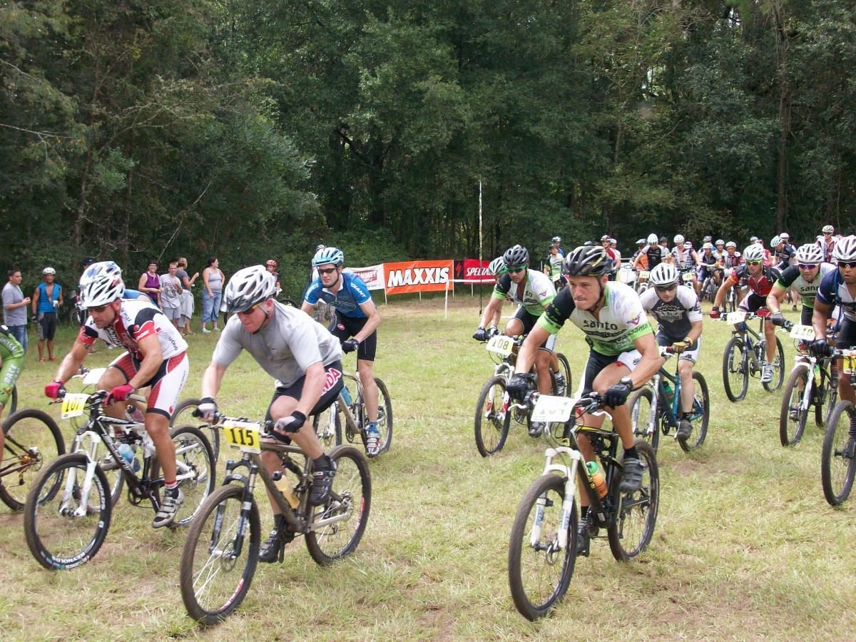 A group of mountain bikers in athletic gear, competing in a cycling race on a grassy terrain. Several riders are in close proximity, with some displaying race numbers on their bikes. Spectators can be seen in the background, watching the event. A large banner with the brand "MAXXIS" is visible, indicating a sponsorship. Haile's Trails mountain bike trail.