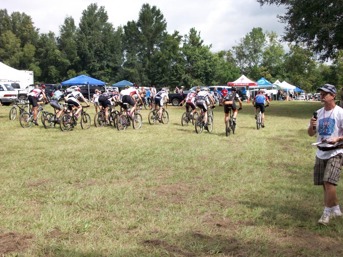 A group of cyclists in athletic gear, riding mountain bikes, are seen from behind as they prepare to start a race in a grassy area. Tents and vehicles are visible in the background, indicating a gathering or event. In the foreground, a man holds a microphone and a clipboard, likely announcing or coordinating the event. Haile's Trails mountain bike trail.