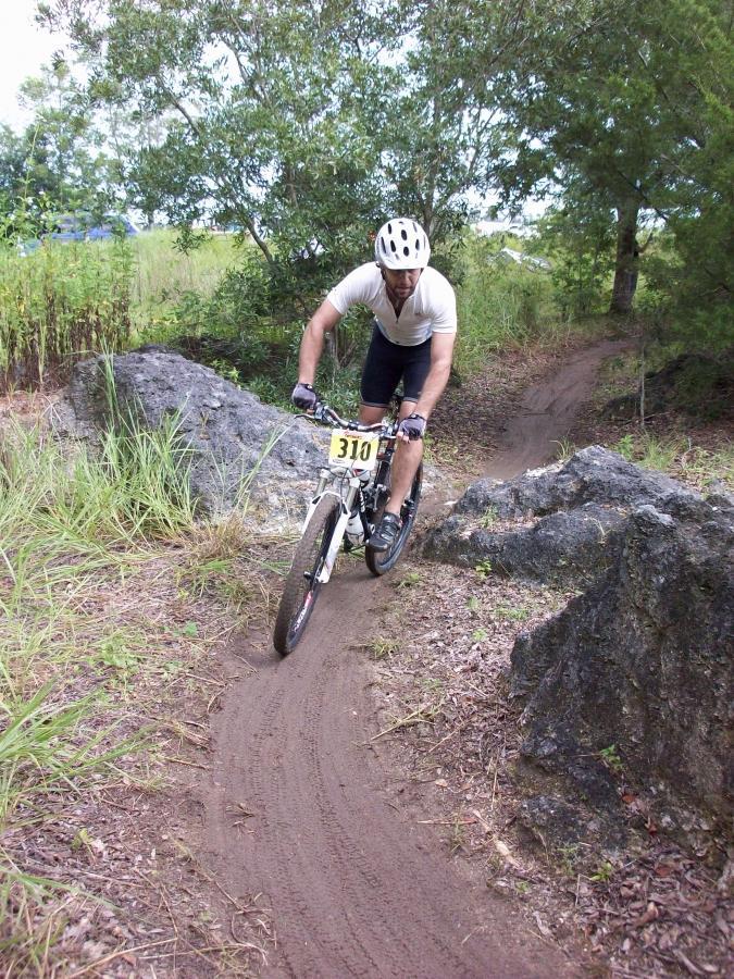 A male mountain biker navigates a winding dirt trail between large rocks, wearing a helmet and a race number. The surrounding area features tall grass and trees in a natural landscape. Haile's Trails mountain bike trail.