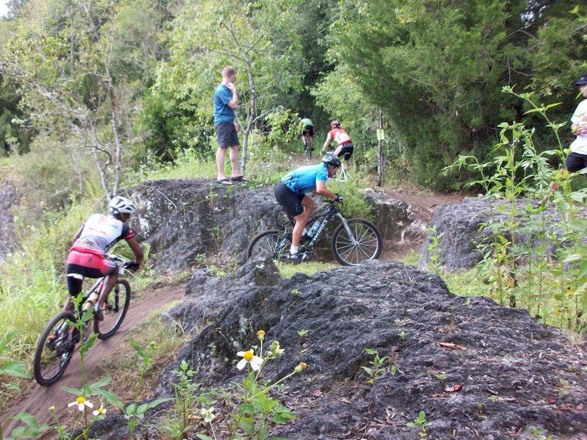 Mountain bikers navigate a rocky trail surrounded by greenery, with one cyclist in a blue jersey and another in a red and black outfit. A few spectators stand in the background, watching the action unfold. Haile's Trails mountain bike trail.