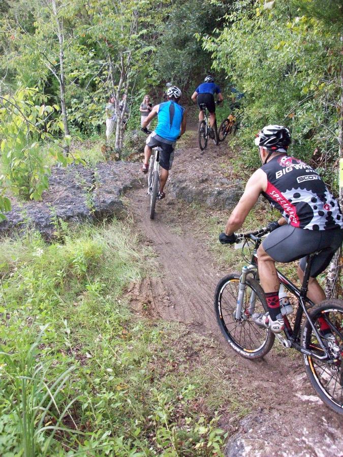 A group of mountain bikers navigating a rocky trail surrounded by greenery. The cyclists, wearing various athletic gear, are riding along a narrow, dirt path lined with grass and trees. Haile's Trails mountain bike trail.