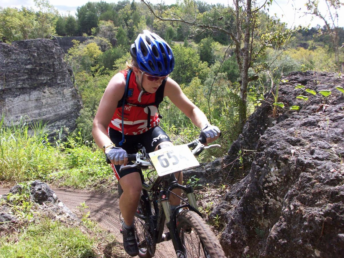 A mountain biker navigates a rocky trail, wearing a blue helmet and a red and white jersey. The background features lush greenery and steep rock formations under a sunny sky. The biker appears focused as they maneuver around obstacles on the path. Haile's Trails mountain bike trail.