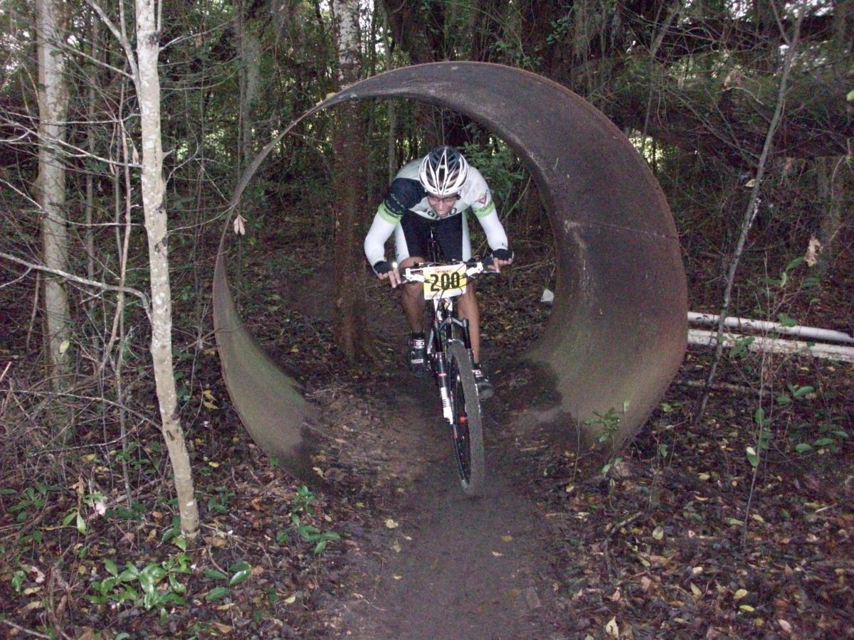 A mountain biker races through a large, circular metal pipe set among trees and foliage in a dense forest. The biker is wearing a helmet and cycling gear, with a race number visible on the front of the bike. The ground is muddy, indicating recent rain, and the surrounding area is rich with greenery. Haile's Trails mountain bike trail.