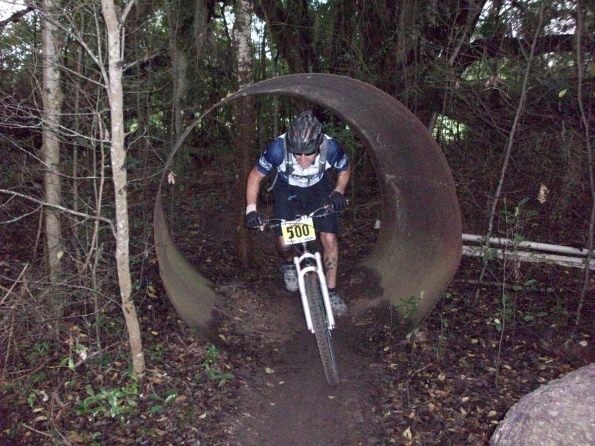A mountain biker wearing a helmet and racing number is navigating through a large metal pipe while riding on a dirt trail in a forested area. Trees and vegetation surround the path, creating a natural outdoor setting. Haile's Trails mountain bike trail.