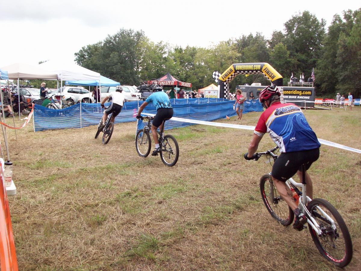 Three mountain bikers race towards the finish line at an outdoor event. In the background, tents and vehicles are set up, and spectators are visible, cheering on the riders. A large finish line banner is displayed prominently. The ground is grassy and slightly uneven, typical of a cycling competition environment. Haile's Trails mountain bike trail.