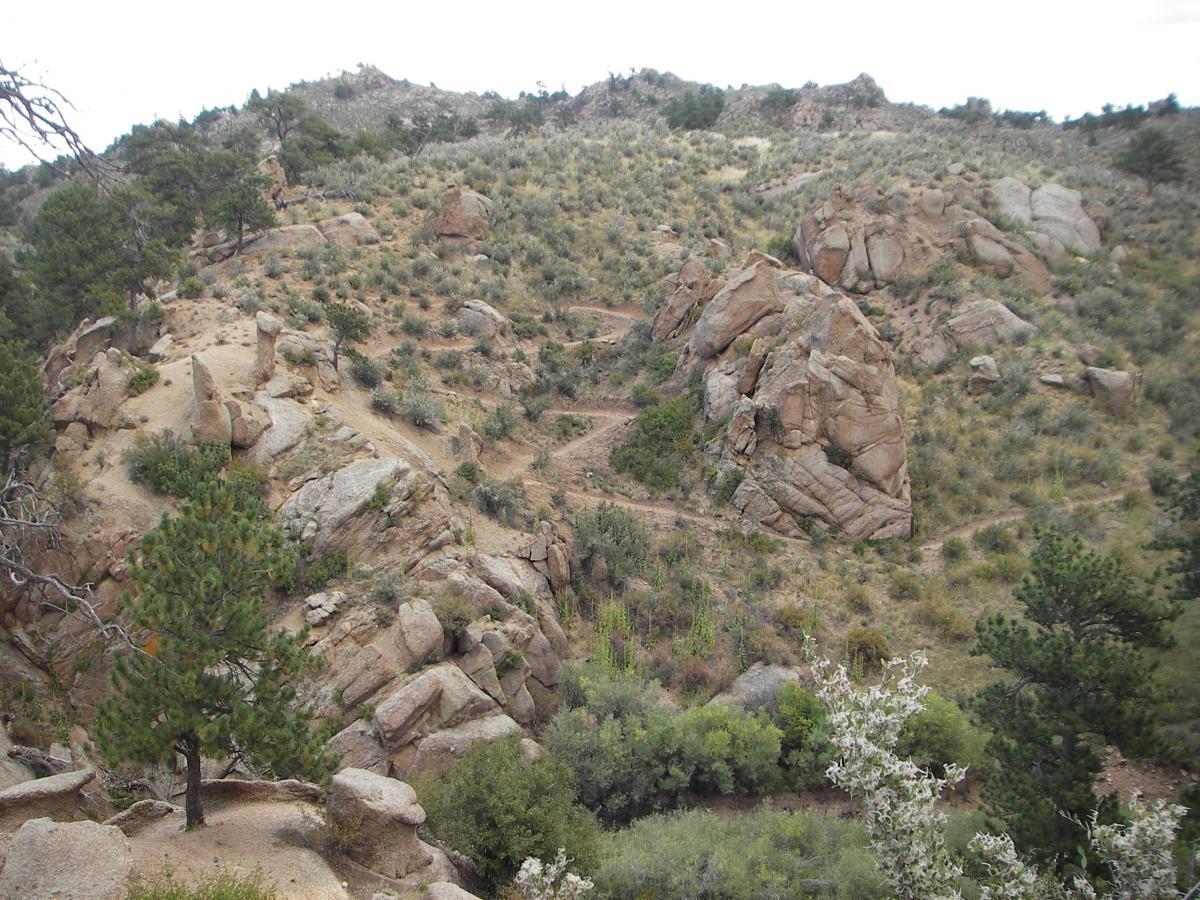A rugged, rocky landscape featuring steep hills and scattered vegetation, including a few trees. The terrain is dry with large boulders and sparse shrubs, indicative of a mountainous area. The sky is overcast, adding a moody ambiance to the scene. Curt Gowdy State Park mountain bike trail.