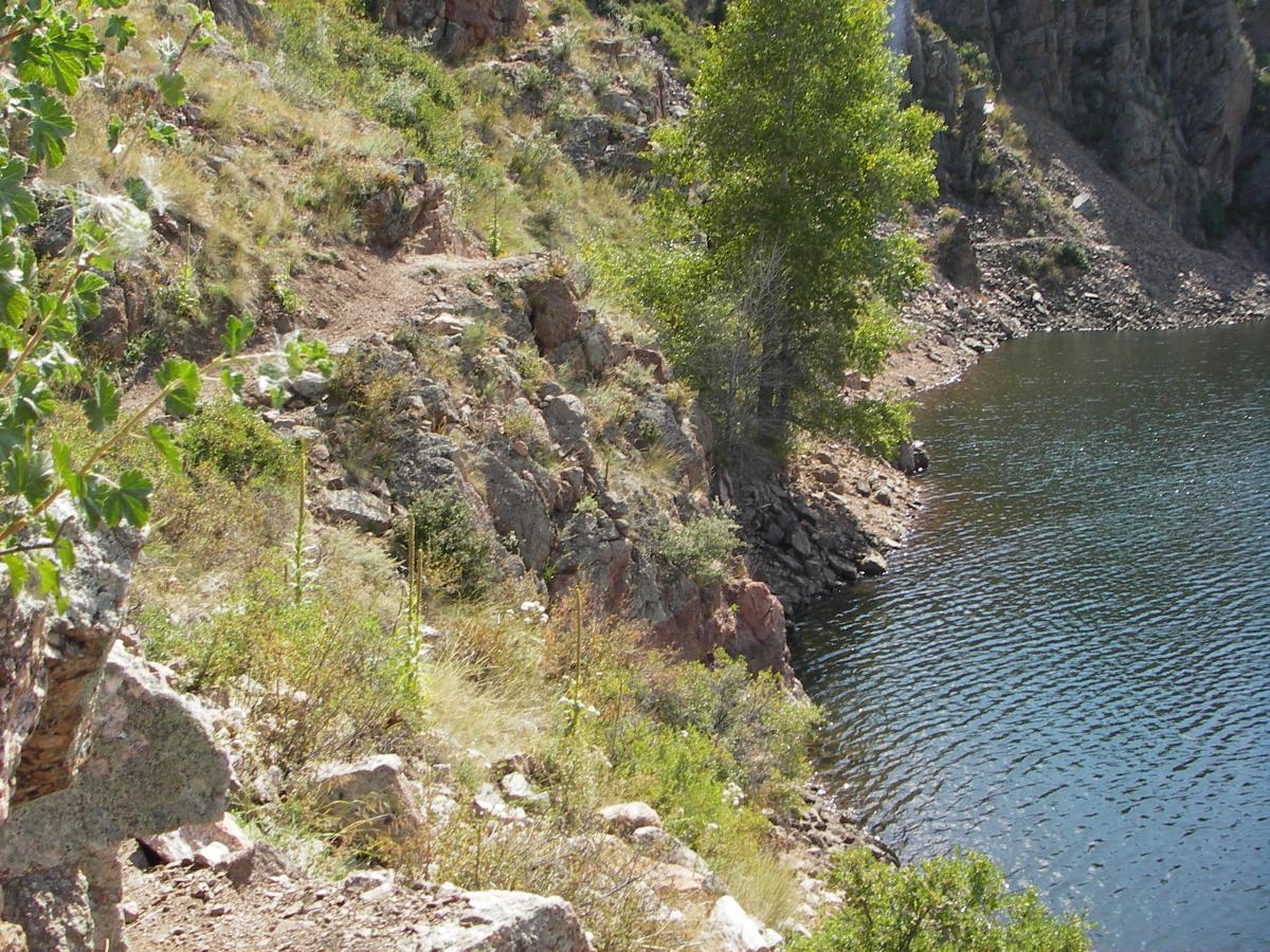 A rocky hillside with sparse vegetation leading to a tranquil body of water, surrounded by steep rocky cliffs and trees. Sunlight casts a warm glow over the scene, highlighting the natural formations and clear water. Curt Gowdy State Park mountain bike trail.