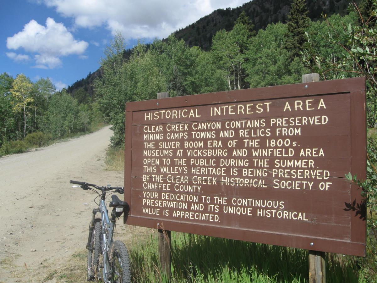 A wooden sign titled "Historical Interest Area" stands beside a dirt path, with a mountain backdrop and green vegetation. The sign describes Clear Creek Canyon's preserved mining camps and relics from the 1800s silver boom era, highlighting the area's historical significance and conservation efforts. A mountain bike is leaned against the sign, suggesting outdoor exploration. CDT: Hope Pass mountain bike trail.