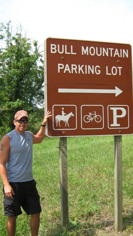 A person stands next to a brown sign that reads "Bull Mountain Parking Lot," pointing to the right. The sign includes icons for horseback riding, biking, and parking. In the background, tall grass and trees are visible under a clear sky. Bull / Jake Mountain mountain bike trail.