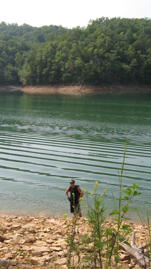 A person standing in shallow water at the edge of a lake, with a rocky shoreline and green trees in the background. The water has gentle ripples, and the scene is bathed in natural light, creating a tranquil atmosphere. Tsali Left Loop mountain bike trail.