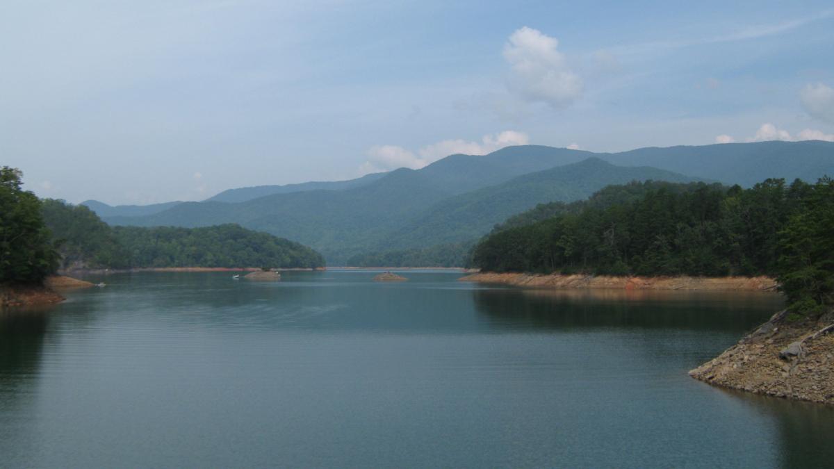 A tranquil lake surrounded by lush green forests and rolling mountains under a partly cloudy sky. The water is calm, reflecting the scenery, and small islands can be seen in the distance. Tsali Left Loop mountain bike trail.