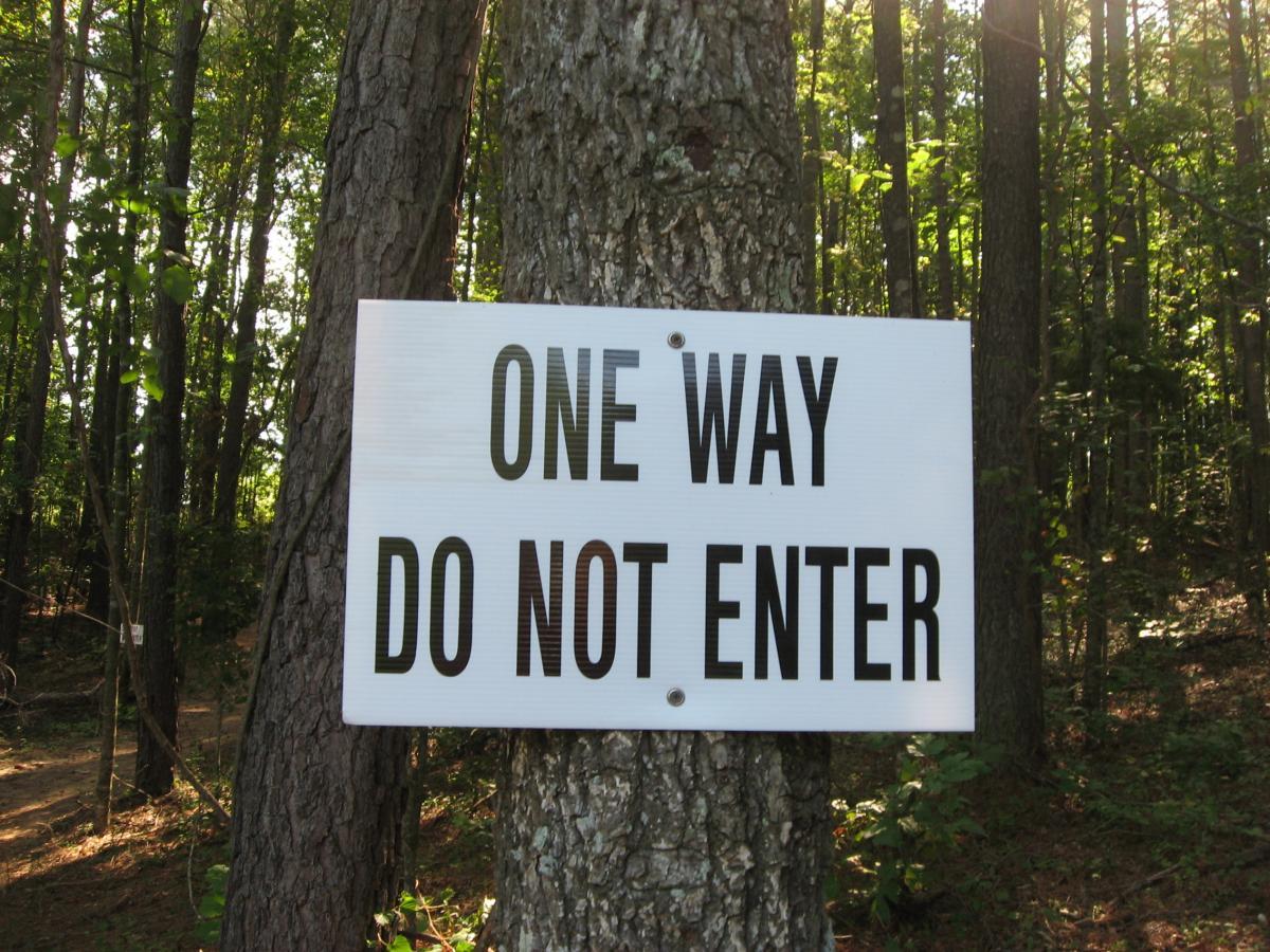 A sign attached to a tree in a wooded area displaying the text "ONE WAY DO NOT ENTER." The background features tall trees and dappled sunlight filtering through the leaves. Harbins Park mountain bike trail.