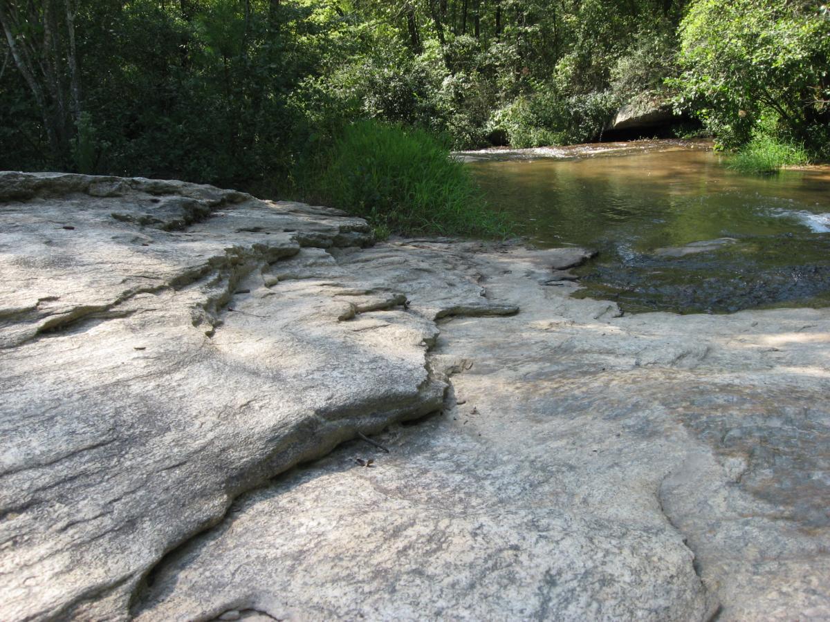 A rocky, natural landscape featuring a smooth stone surface leading down to a gently flowing stream, surrounded by lush green vegetation and trees. The scene captures the tranquility of nature with dappled sunlight filtering through the foliage. Harbins Park mountain bike trail.
