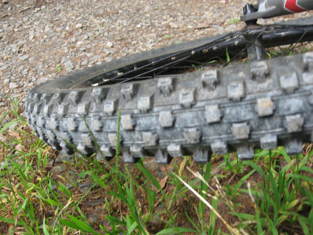 A close-up view of a mountain bike tire resting on a gravel and grass surface, showcasing the textured tread pattern designed for traction on uneven terrain. Stanley Gap mountain bike trail.