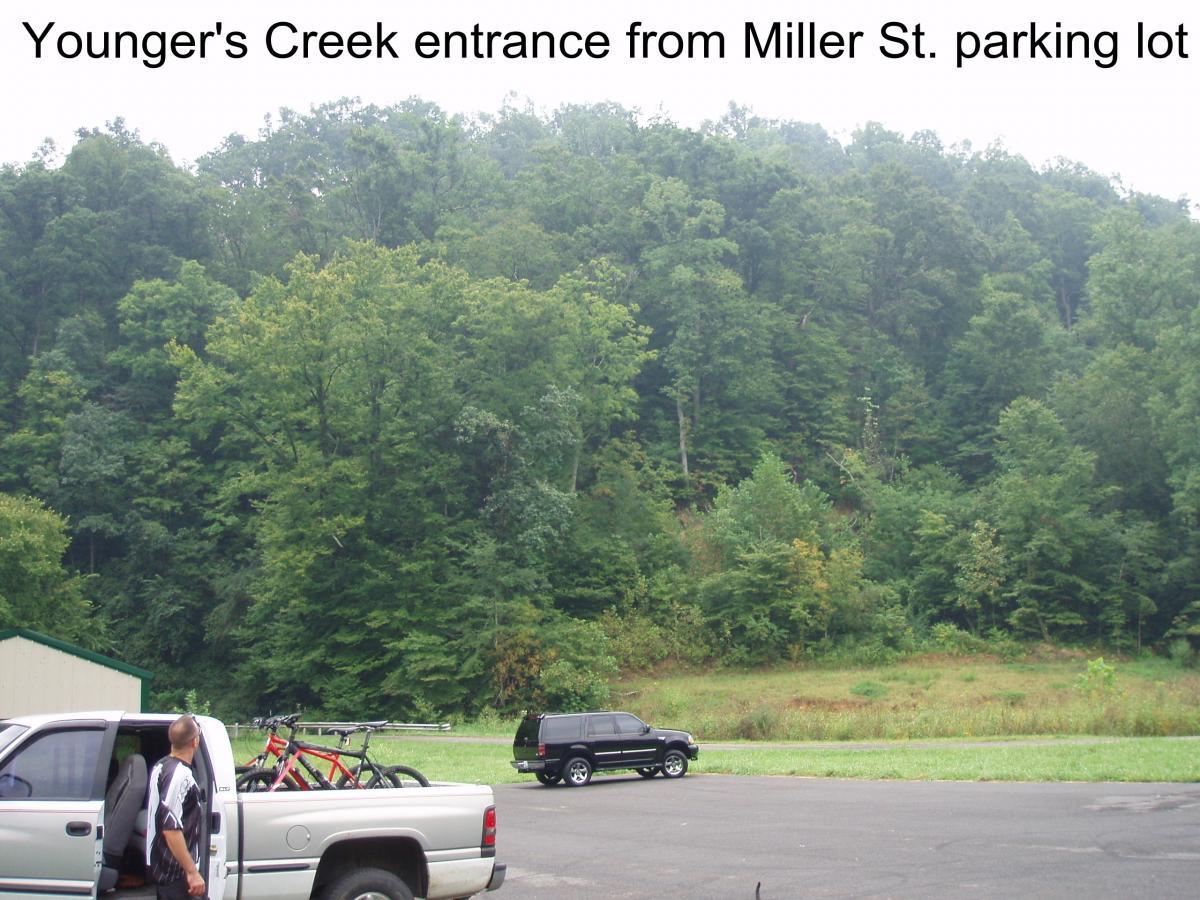 Alt text: A view of the entrance to Younger’s Creek from the Miller Street parking lot, featuring a lush green hillside, two parked vehicles (one silver truck with bikes in the bed and one black SUV), and a person standing near the truck looking towards the trees. Youngers Creek mountain bike trail.