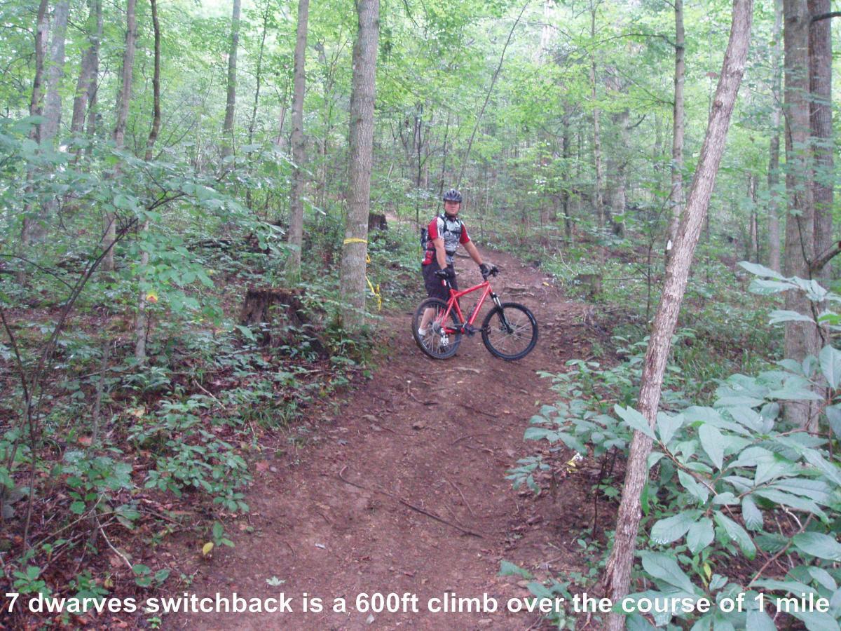 A mountain biker pauses on a winding dirt trail surrounded by lush green trees in a forest. The trail, known as "7 Dwarves Switchback," features a steep incline and is marked as a challenging 600-foot climb over one mile. Youngers Creek mountain bike trail.