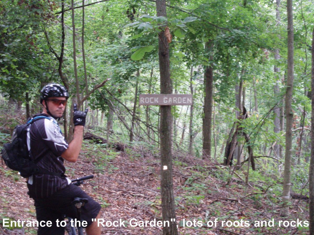 Mountain biker at the entrance to the "Rock Garden" trail, surrounded by dense greenery and trees, with a sign indicating the trail name. The biker is wearing a helmet and gloves, looking back while giving a thumbs-up gesture. Youngers Creek mountain bike trail.