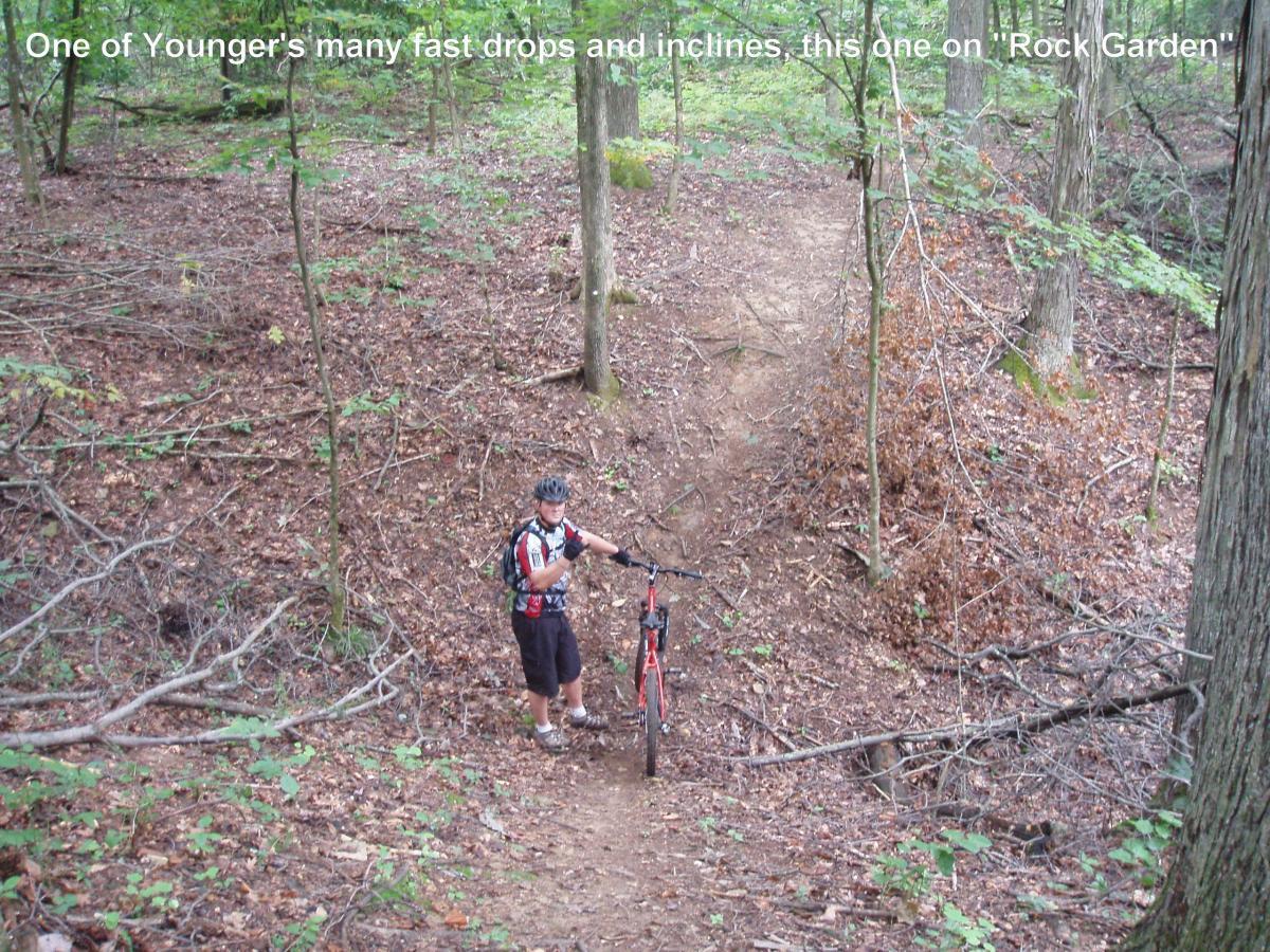 A mountain biker standing next to his bike on a trail in a wooded area, surrounded by fallen leaves and trees. The path includes a steep incline leading up through the forest. Youngers Creek mountain bike trail.