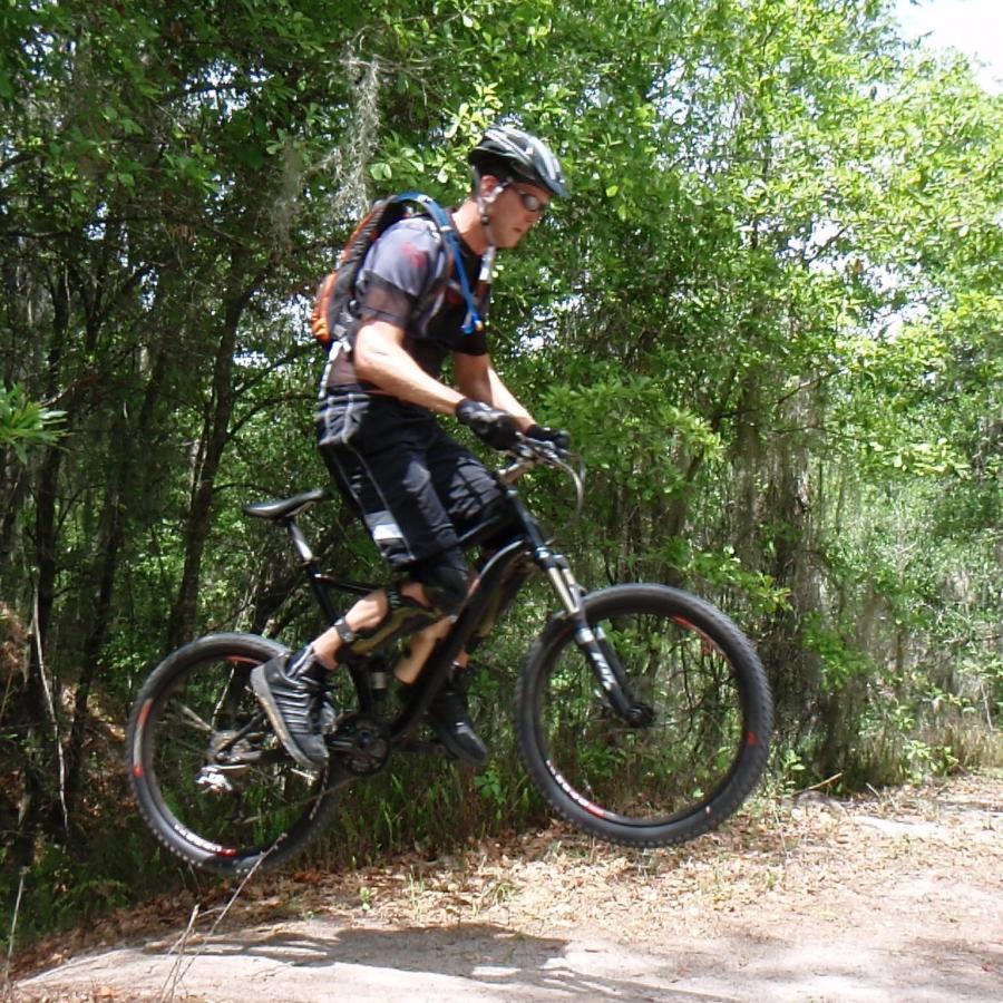 A cyclist performing a jump on a mountain bike in a forested area, wearing a helmet and protective gear. Lush greenery surrounds the dirt trail, with sunlight filtering through the trees. Alafia River State Park mountain bike trail.