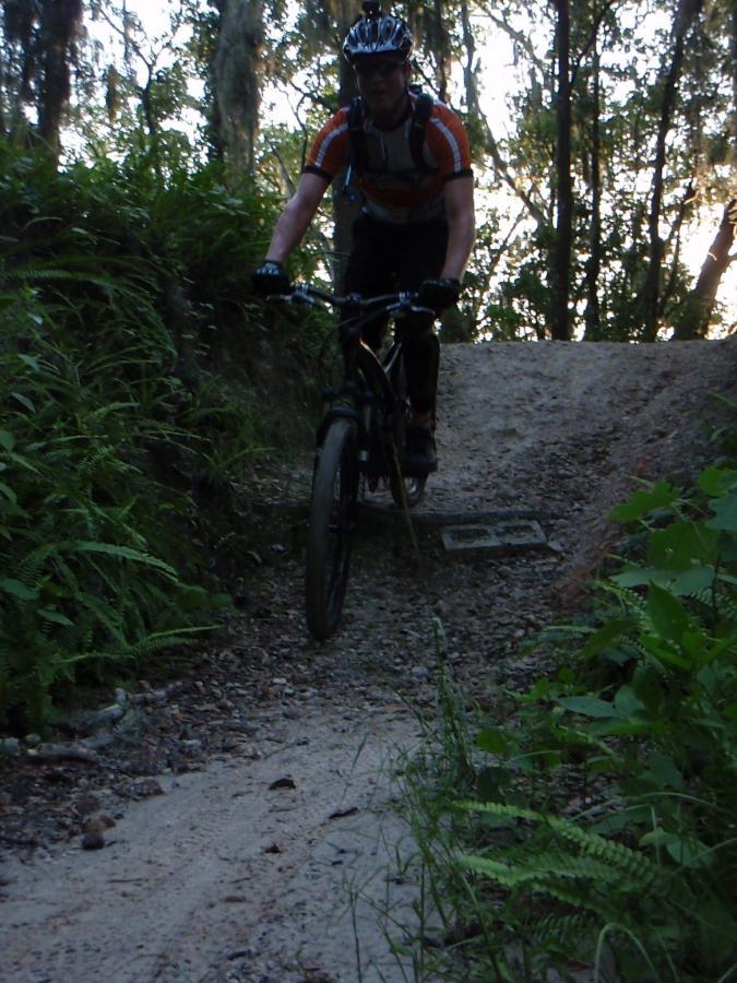 A mountain biker pedaling along a gravel trail surrounded by lush greenery and tall trees in a natural setting. The rider, wearing a helmet and colorful sports attire, is focused on navigating the uneven terrain. Loyce E. Harpe Park mountain bike trail.