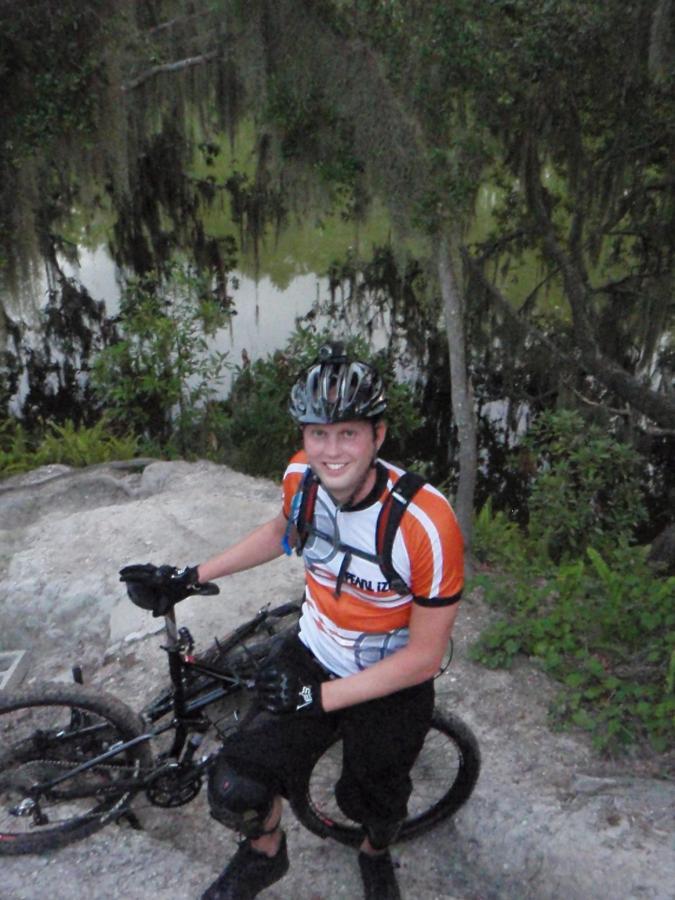 A smiling young man in a biking outfit sits next to a mountain bike, with a scenic background of trees and water. He is wearing a helmet and protective gear, displaying a thumbs-up gesture, indicating enjoyment of the outdoor activity. Loyce E. Harpe Park mountain bike trail.