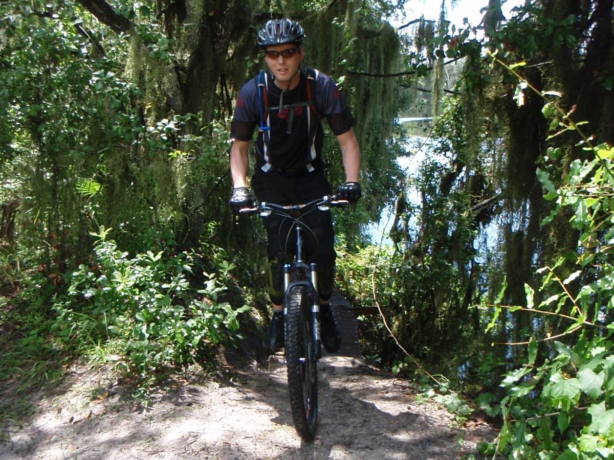 A mountain biker navigating a narrow trail surrounded by lush greenery and trees, with a body of water visible in the background. The cyclist is wearing a helmet, sunglasses, and protective gear while riding a mountain bike. Loyce E. Harpe Park mountain bike trail.