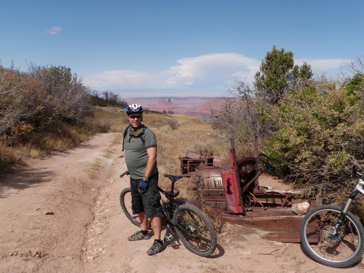 A person wearing a helmet stands on a dirt path surrounded by dry grass and sparse vegetation, with a vintage, rusted vehicle partially hidden in the bushes beside them. In the background, a scenic view of a canyon and blue sky with some clouds is visible, while two mountain bikes rest on the ground nearby. The Whole Enchilada mountain bike trail.