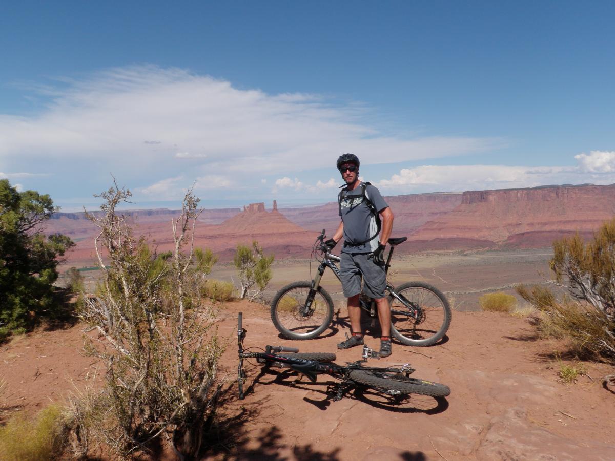 A person standing next to a mountain bike on a rocky outcrop, overlooking a vast desert landscape with red rock formations and blue skies. The individual is wearing casual cycling attire and a helmet, with another bike lying on the ground nearby. Vegetation is sparse, typical of a desert environment. The Whole Enchilada mountain bike trail.