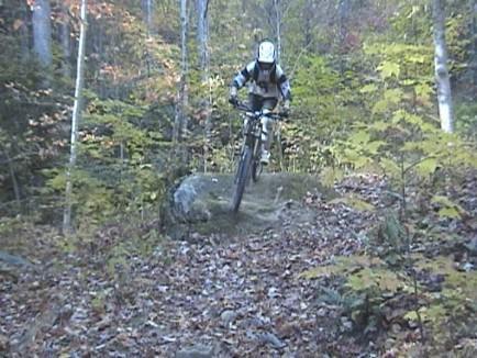 A mountain biker in protective gear jumps off a rock on a forested trail surrounded by autumn foliage. Table Rock mountain bike trail.