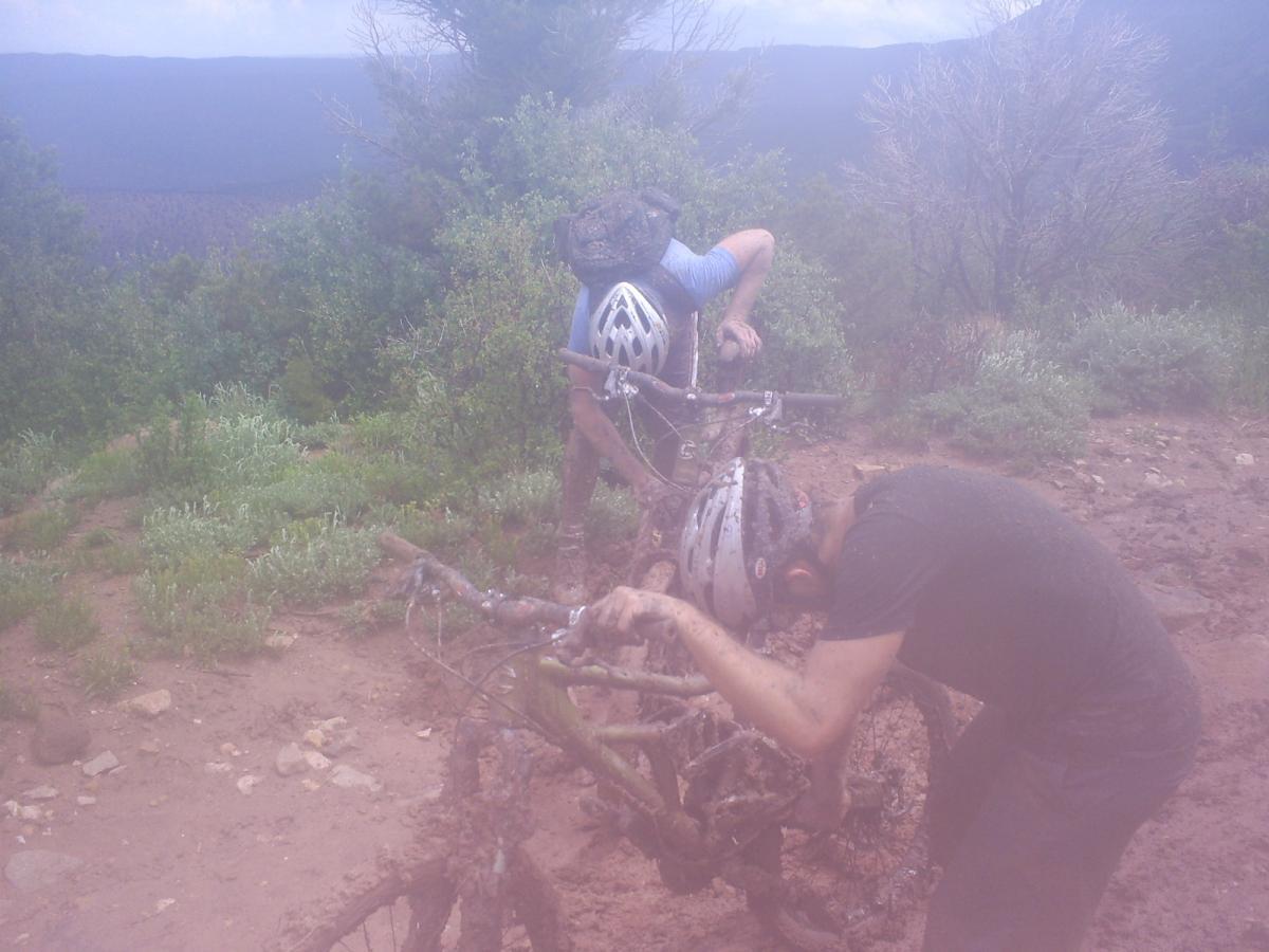 Two mountain bikers work to clean their muddy bikes after a ride on a rugged trail. The background features green foliage and a mountainous landscape under a cloudy sky. The bikes are caked in mud, and the riders appear focused and engaged in their task. The Whole Enchilada mountain bike trail.