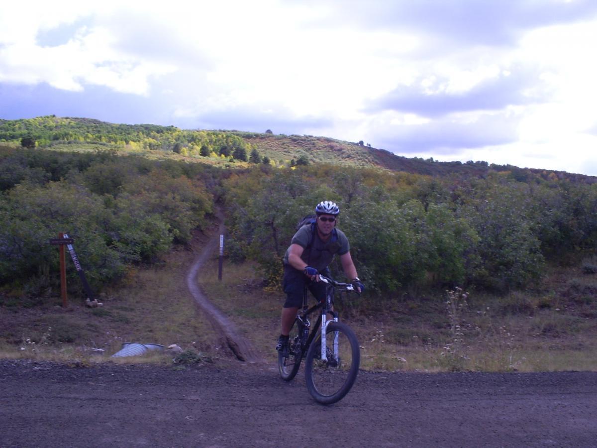 A mountain biker riding along a dirt path surrounded by greenery and rolling hills, with a partly cloudy sky overhead. The biker is wearing a helmet and sunglasses, and there are trail signs visible in the background. The Whole Enchilada mountain bike trail.