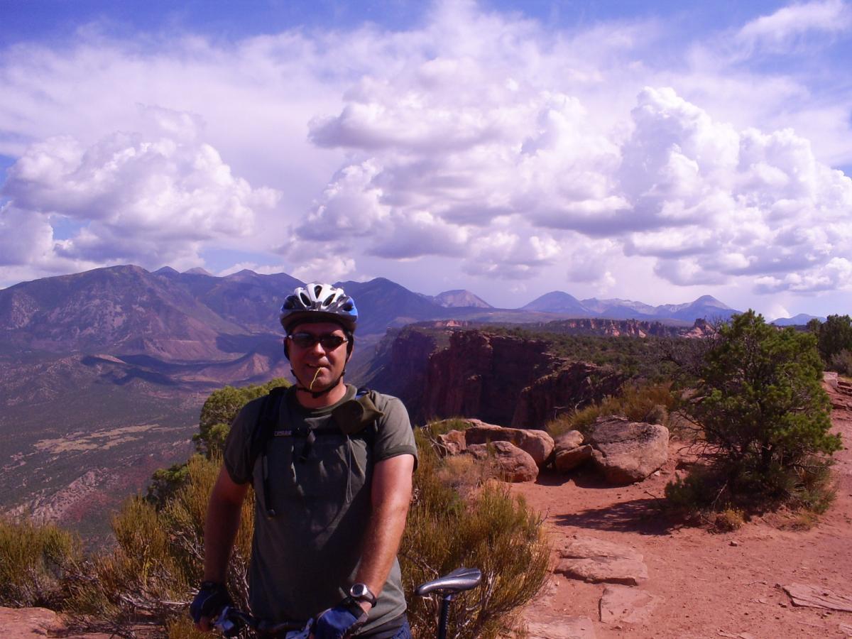 A person wearing a helmet and sunglasses stands beside a mountain bike, with a panoramic view of rugged mountains and a cloudy sky in the background. The landscape features rocky terrain and sparse vegetation, indicating a scenic outdoor cycling location. The Whole Enchilada mountain bike trail.