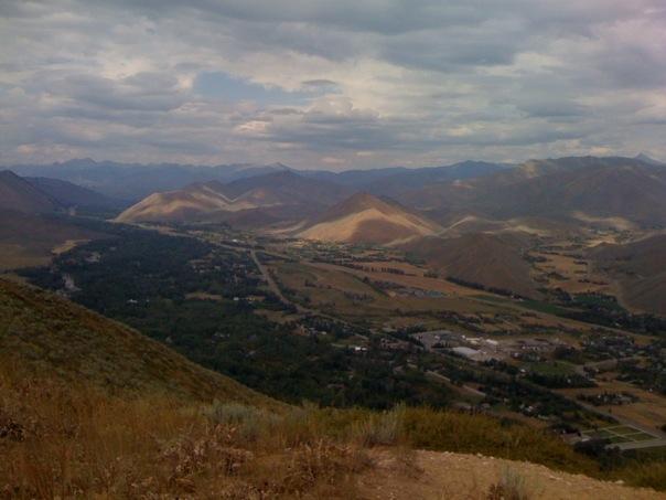 A panoramic view of a valley surrounded by hills and mountains, under a cloudy sky. The foreground features grassy terrain leading to the slopes, while the background showcases undulating hills with varying shades of brown and green. A small town is visible at the bottom of the valley, dotted with houses and fields. Carbonate Training Loop mountain bike trail.