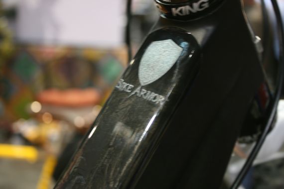 Close-up image of a bicycle frame featuring the engraving "State Armor" on the top tube, with a blurred colorful background that includes abstract patterns. The bike components are partially visible, highlighting the detail in the frame's design.