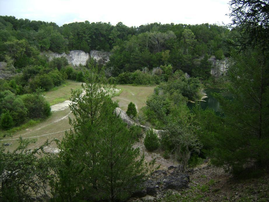 A serene landscape featuring a natural quarry surrounded by lush greenery and trees. The scene includes rocky cliffs in the background, a grassy area in the foreground, and a calm body of water at the edge, under a cloudy sky. Haile's Trails mountain bike trail.