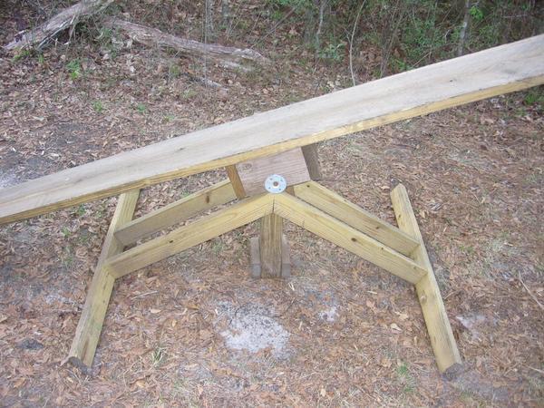 A wooden seesaw positioned on the ground in a natural setting, surrounded by fallen leaves and trees. The seesaw features a long wooden plank balanced on a central pivot, supported by a triangular wooden frame. The Rock Trail mountain bike trail.