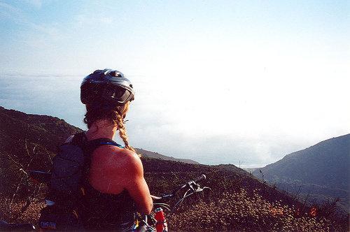 A cyclist with braided hair wearing a helmet, looking out over a scenic landscape with mountains and a foggy ocean view in the distance. The scene captures a sense of adventure and tranquility. Sycamore Canyon mountain bike trail.