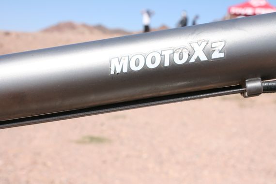 Close-up of a silver bike frame with the brand name "MOOTOxZ" prominently displayed. The background features a desert landscape with blurred figures in the distance, suggesting an outdoor activity or event.